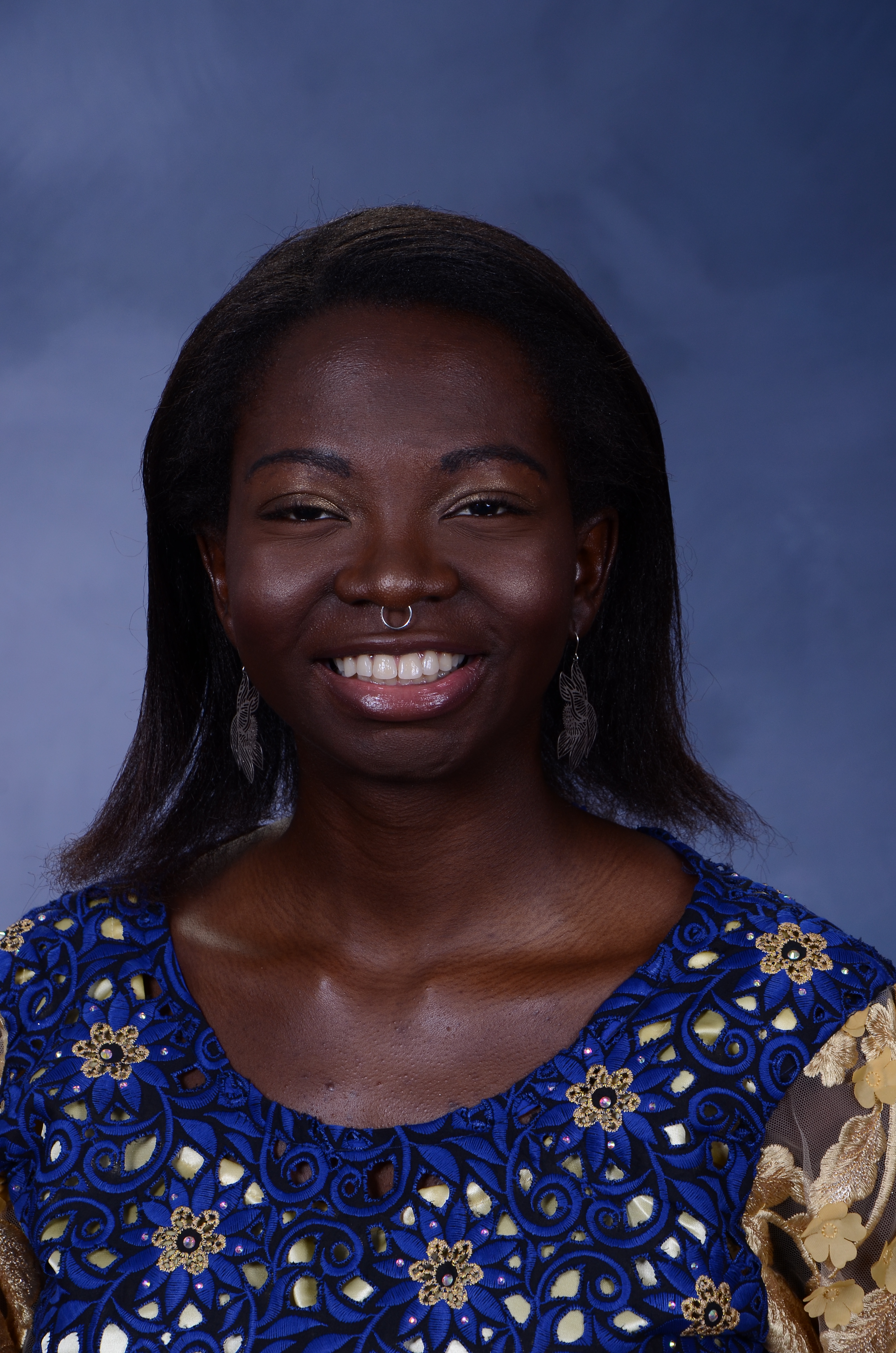 Aderonke - a person with dark brown skin, shoulder length brown hair, a silver septum ring, and braces - wears a white dress, smiles, and stands in front of Sterling Memorial Library at Yale University.