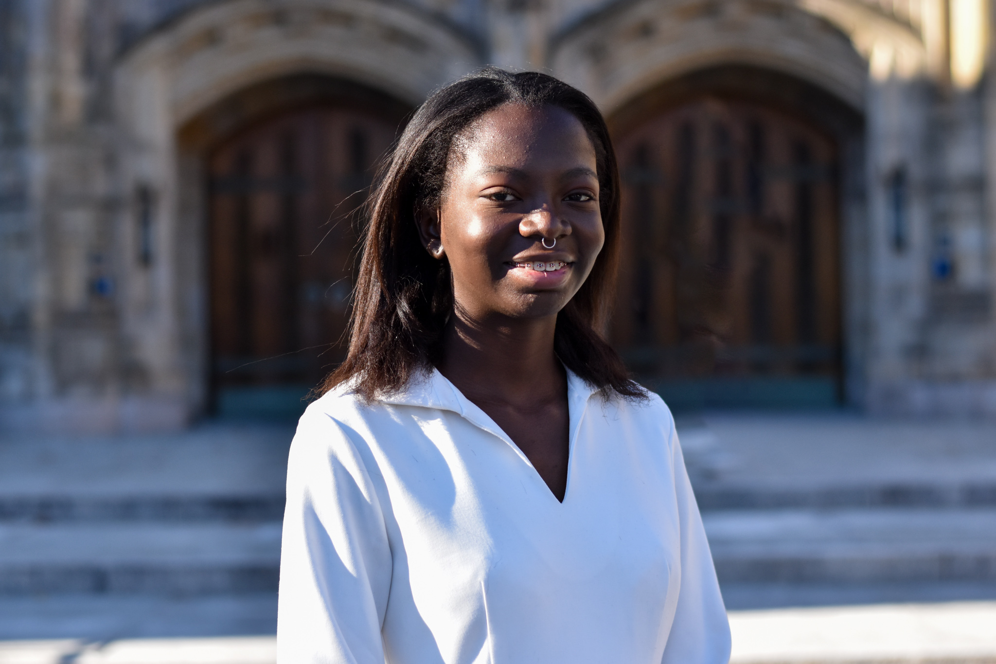 Aderonke - a person with dark brown skin, shoulder length brown hair, a silver septum ring, and braces - wears a white dress, smiles, and stands in front of Sterling Memorial Library at Yale University.